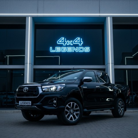 A sleek black Toyota Hilux parked in front of a modern building with a neon blue '4x4 Legends' sign.